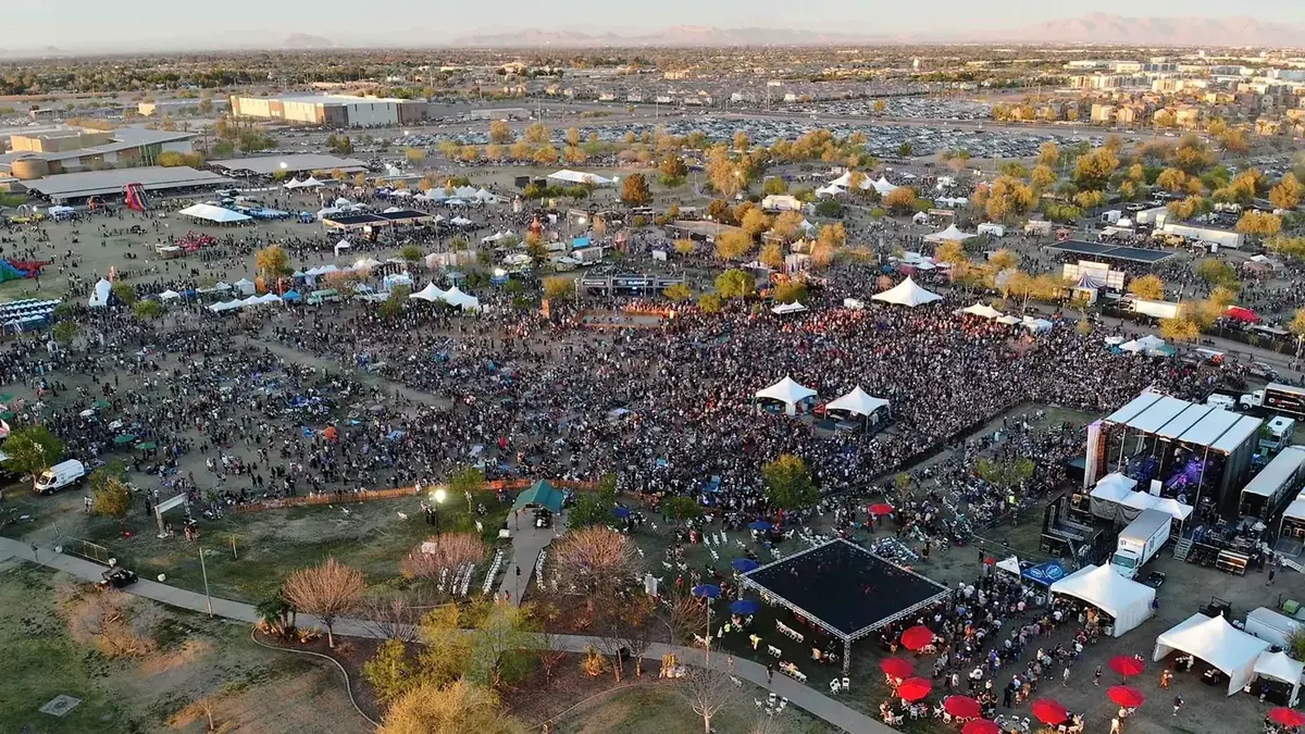 Aerial view of the festival at dusk: a dense crowd fills the grounds in front of the main stage, ringed by white vendor tents, food trucks, a dance floor and a packed parking lot, with the valley and mountains on the horizon.