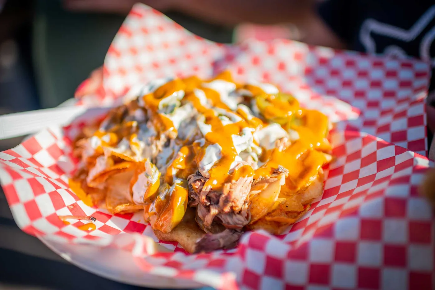 A basket of BBQ nachos in red-and-white checkered paper: tortilla chips piled with pulled pork, melted cheese sauce, a sour cream drizzle and sliced pickled peppers.