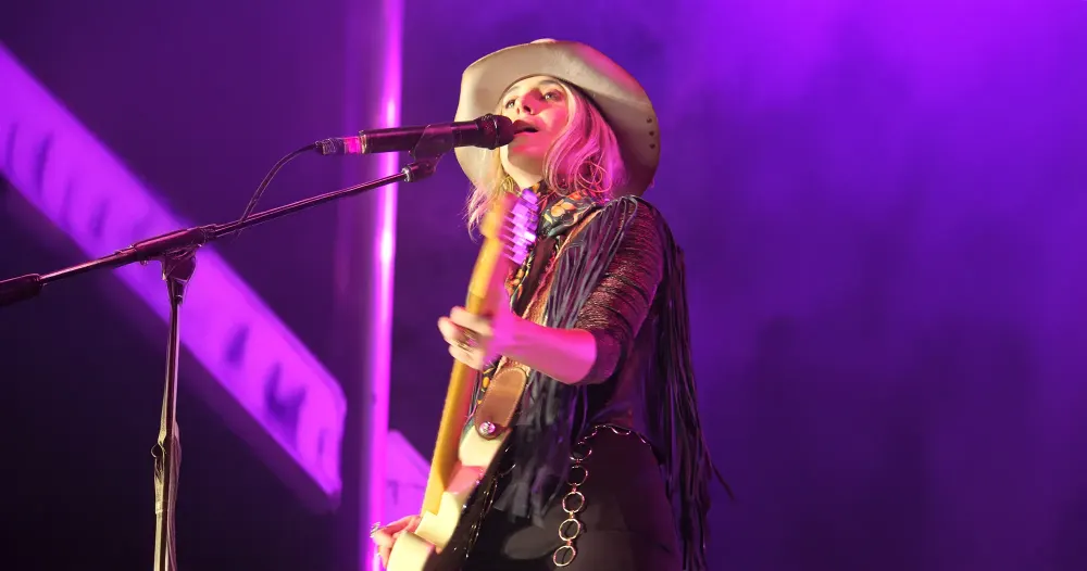 A performer in a wide-brimmed hat sings into the microphone while playing guitar under purple stage lights at a past Desert Sky Music Festival.